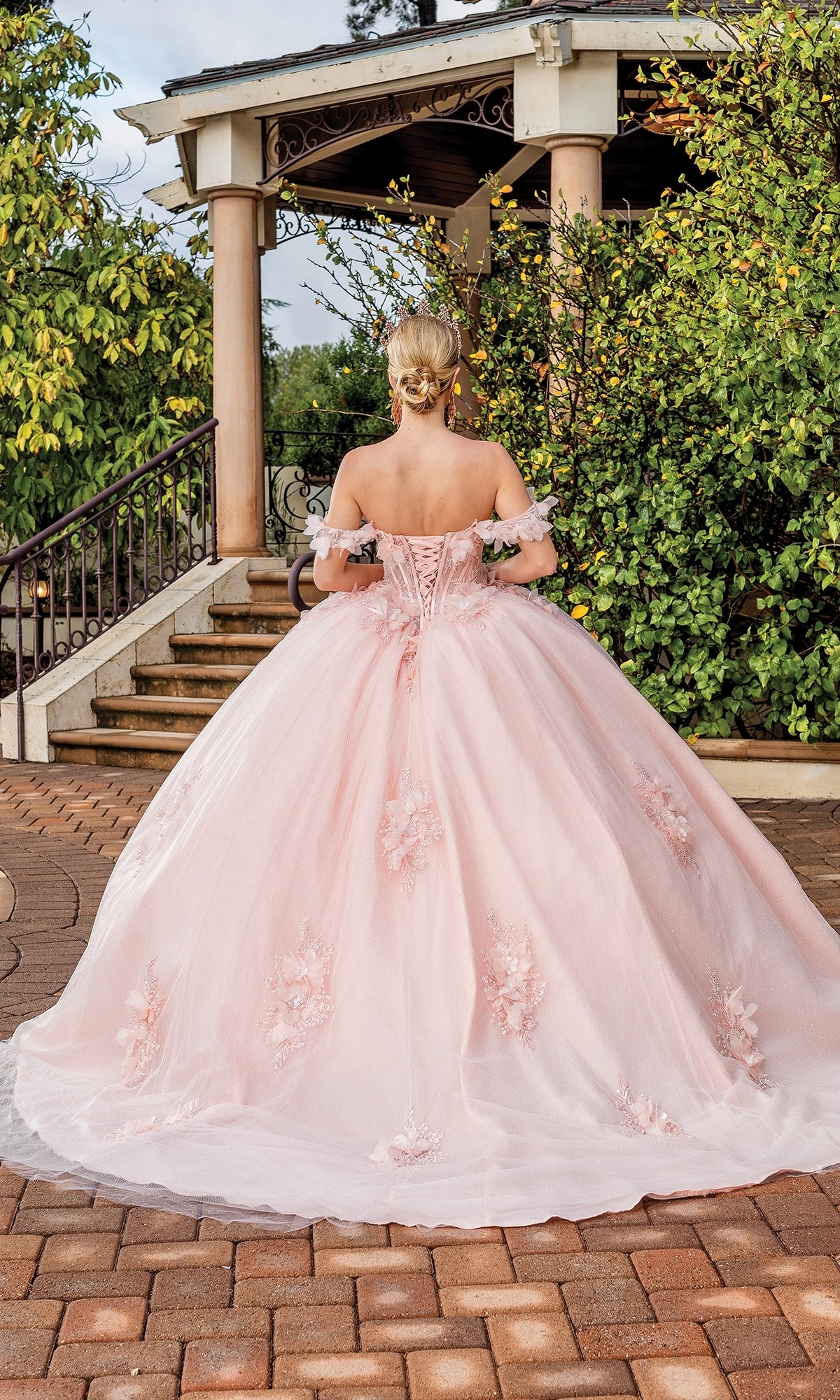 A person in the Quinceanera Dress 1883 by Dancing Queen, an off-the-shoulder pink ball gown with floral details, stands outdoors near stairs and a gazebo surrounded by greenery.